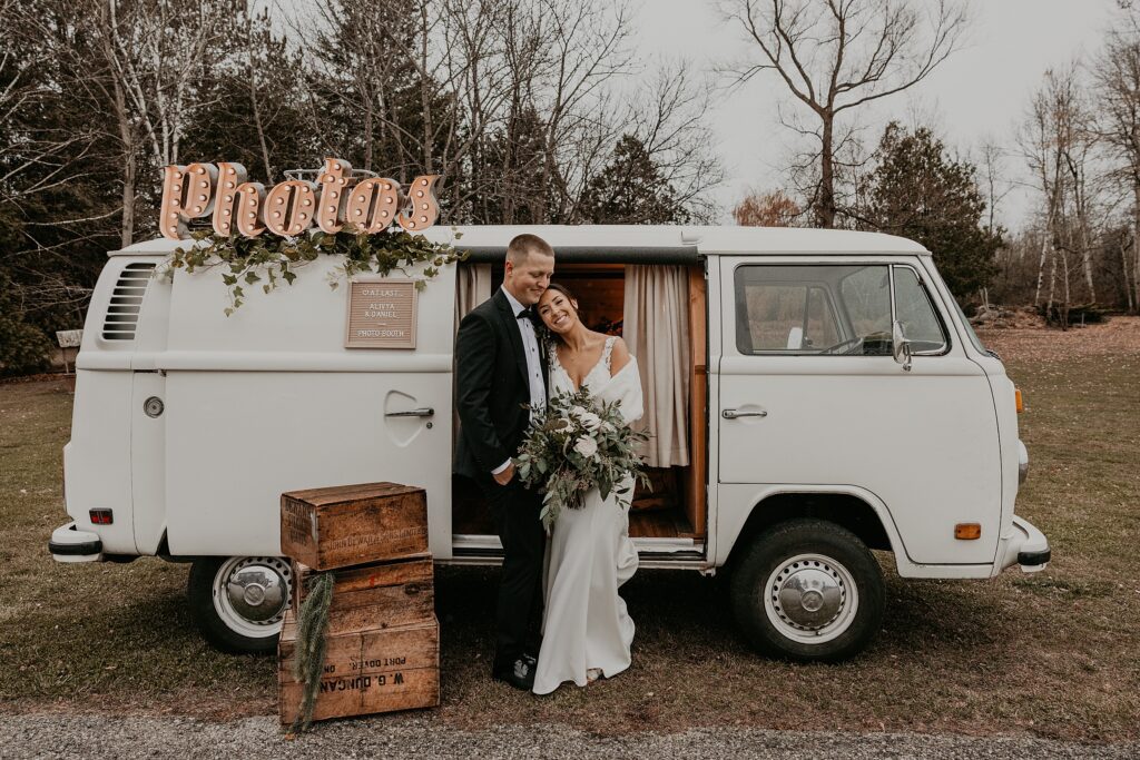 Bride and groom cozy up to each other in front of a vintage white VW bus converted into a photobooth. The bride is smiling at the camera and resting her head on the groom's shoulder. The groom's head is leaning into the bride's and looking down. Captured by best newmarket wedding photographer ashlee ellison.