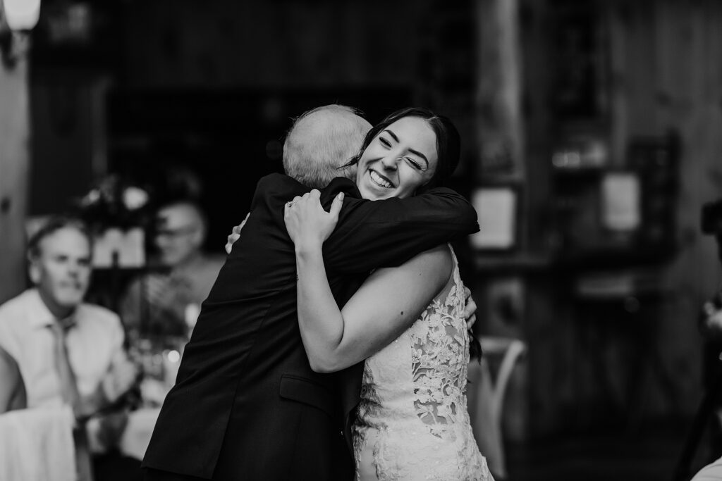 Black and white image of joyful bride sharing a hug with her father at her holland marsh wineries wedding reception. captured by top newmarket wedding photographer ashlee ellison.