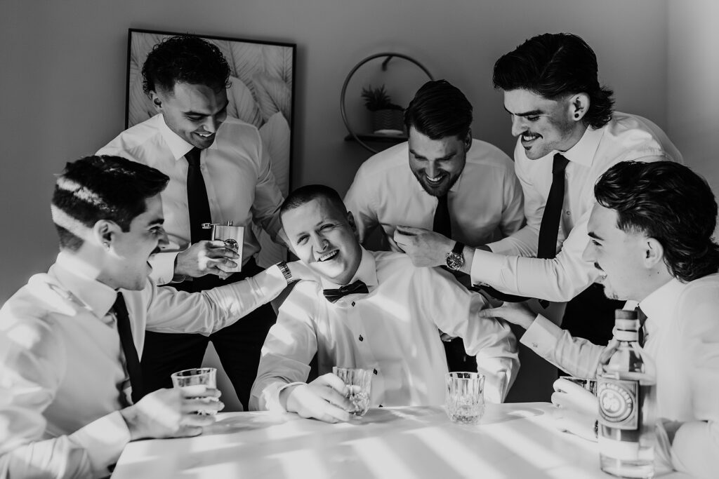 After getting ready for his wedding, a groom is sitting with his groomsmen at a table drinking a bottle of Wayne Gretzky spirits. Three groomsmen are standing behind him, two are sitting at the table with him. All are looking at the groom, holding their classes, they have their hand on the grooms shoulder or back. All are laughing. Captured by best wedding photographer in ontario, canada, ashlee ellison.