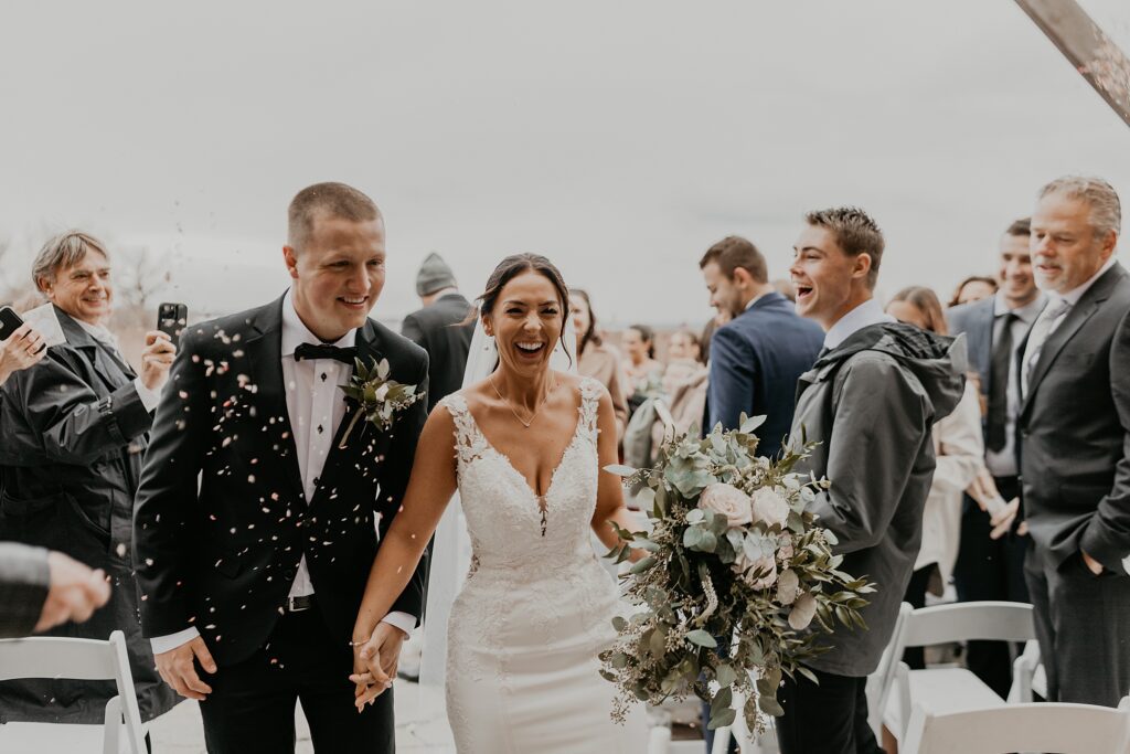 Bride and groom hold hands and laugh as they walk back up the aisle at their Holland Marsh Wineries wedding. Guests look on as they throw white confetti in the air. Captured by best newmarket wedding photographer Ashlee Ellison.