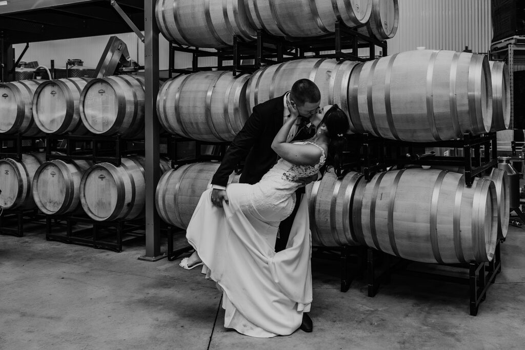 black and white image. bride and groom share a kiss in the cellar at the holland marsh wineries wedding. groom is dipping the bride and her leg is extended. the bride's hand is resting on the groom's neck. captured by best newmarket wedding photographer ashlee ellison.