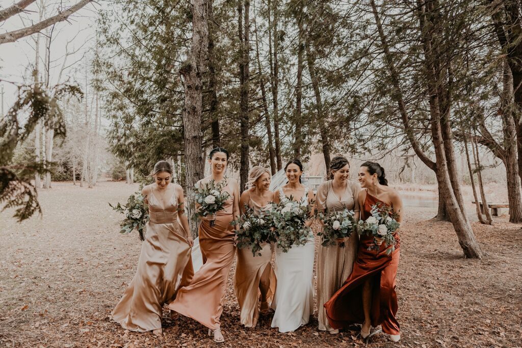 Bride and bridesmaids in champagne coloured gowns are walking amongst trees at holland marsh wineries in newmarket, ON. the girls are smiling and laughing. captured by best newmarket wedding photographer ashlee ellison.
