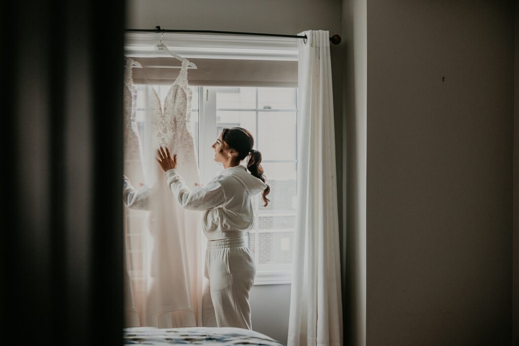 A bride is standing in front of a window looking at her gown hanging atop the curtain rod. She is touching the front of it. Captured by top newmarket wedding photographer ashlee ellison.
