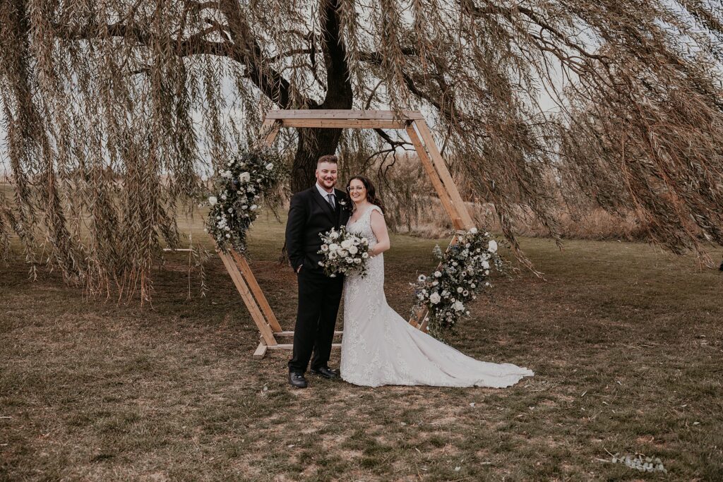 Bride and groom pose for a wedding day portrait in front of their wedding hexagonal wood wedding altar adorned with anemone bouquets. Bride and groom are smiling at the camera. captured by top sarnia wedding photographer.