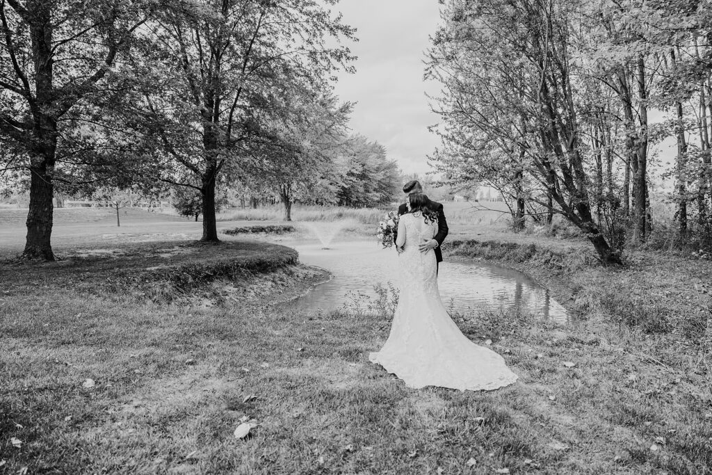 Black and white image of a bride and groom sharing a cuddle in front of a pond on the grounds of Crescent Hill Acres.