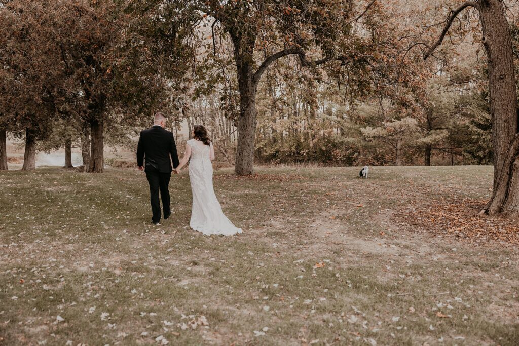 Bride and groom are walking on a grassy area at crescent hill acres. Their backs are to the camera. Their dog is walking alongside of them.