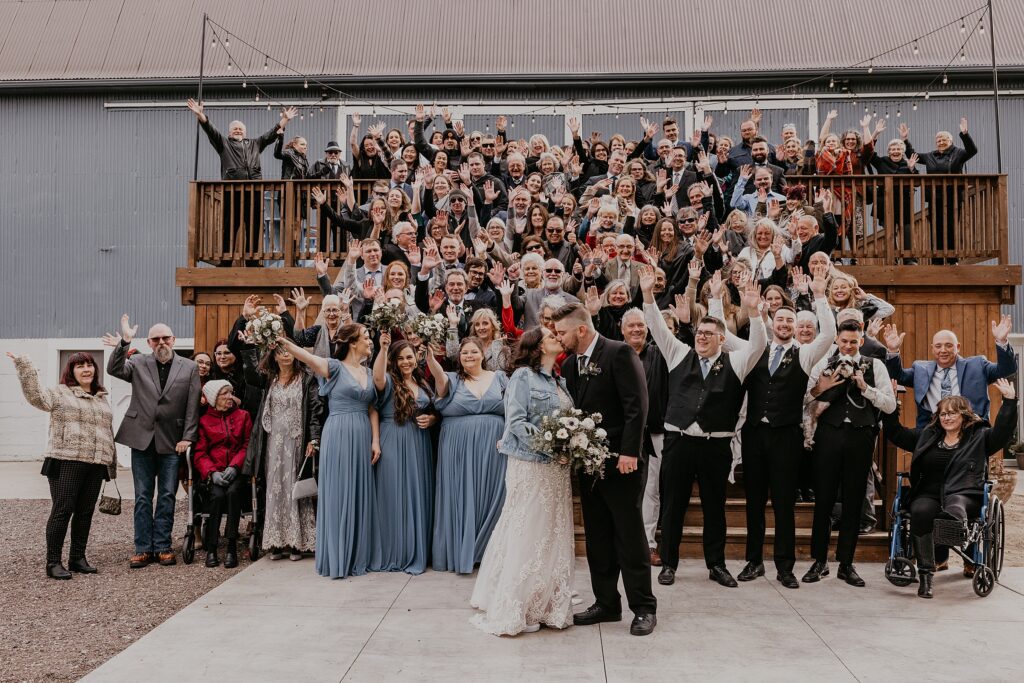 Image of all wedding guests on the stairs of Crescent Hill Acres. The wedding party and guests have their arms thrown up in celebration while the bride and groom kiss in front of them. Captured by top sarnia wedding photographer ashlee ellison.