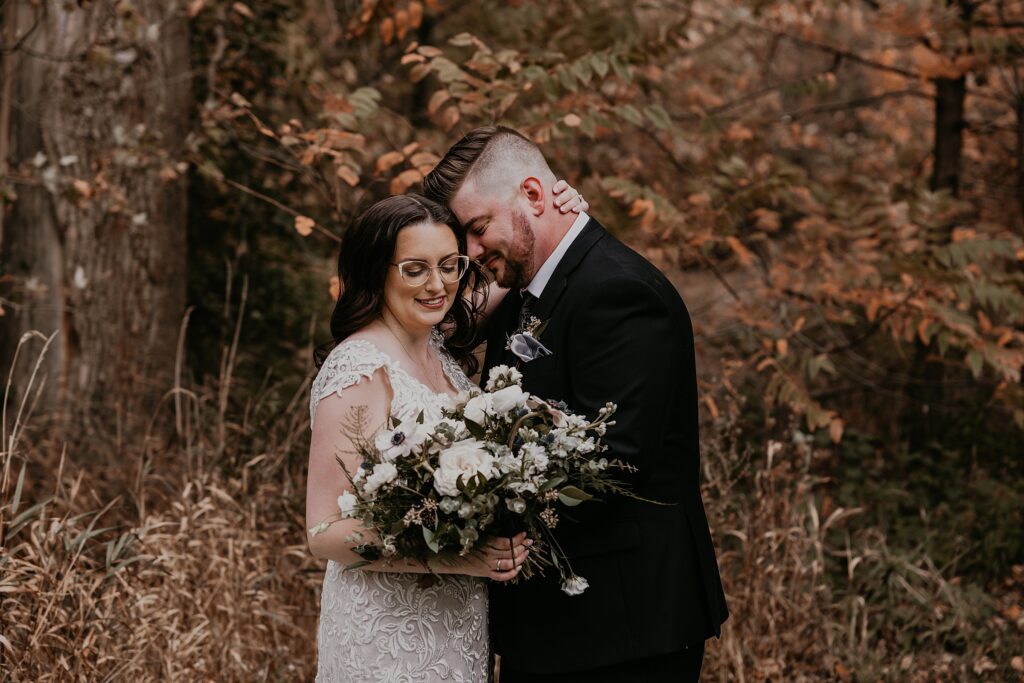 Bride and groom pose for an intimate wedding portrait in a forested area of Crescent Hill Acres. the groom is resting his forehead against his brides temple. the brides eyes are closed and looking down at her bouquet of anemones. captured by top sarnia wedding photographer ashlee ellison.