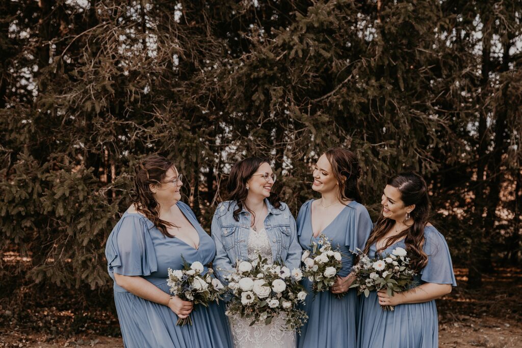 Bride and bridesmaids are standing for a casual portrait at Crescent Hill Acres. Bridesmaids are wearing blue gowns and the bride's lace gown is covered with a denim jacket. They are all holding bouquets of anemones and laughing while looking at each other. Captured by top sarnia wedding photographer ashlee ellison.