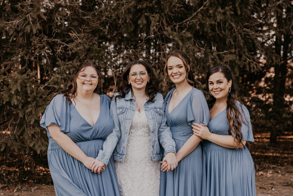 Bridesmaids in long blue gowns are standing with the bride in a white lace gown and denim jacket posing for a casual portrait at Crescent Hill Acres. They are all holding hands and smiling at the camera. Captured by top sarnia wedding photographer ashlee ellison.