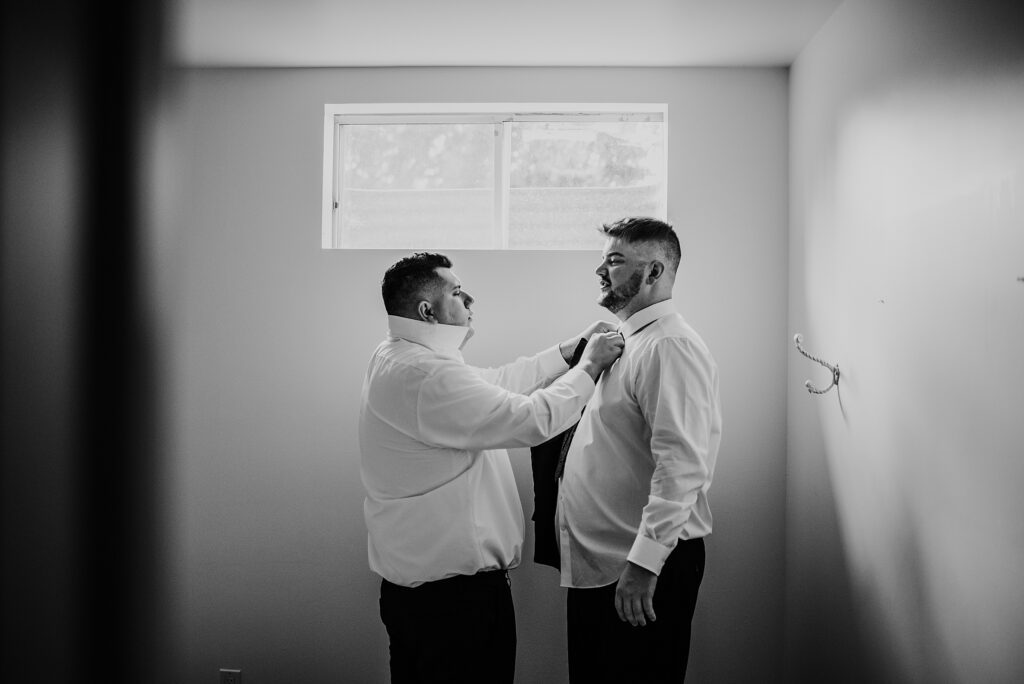 Groomsman is helping the groom adjust his tie while getting ready for his wedding.