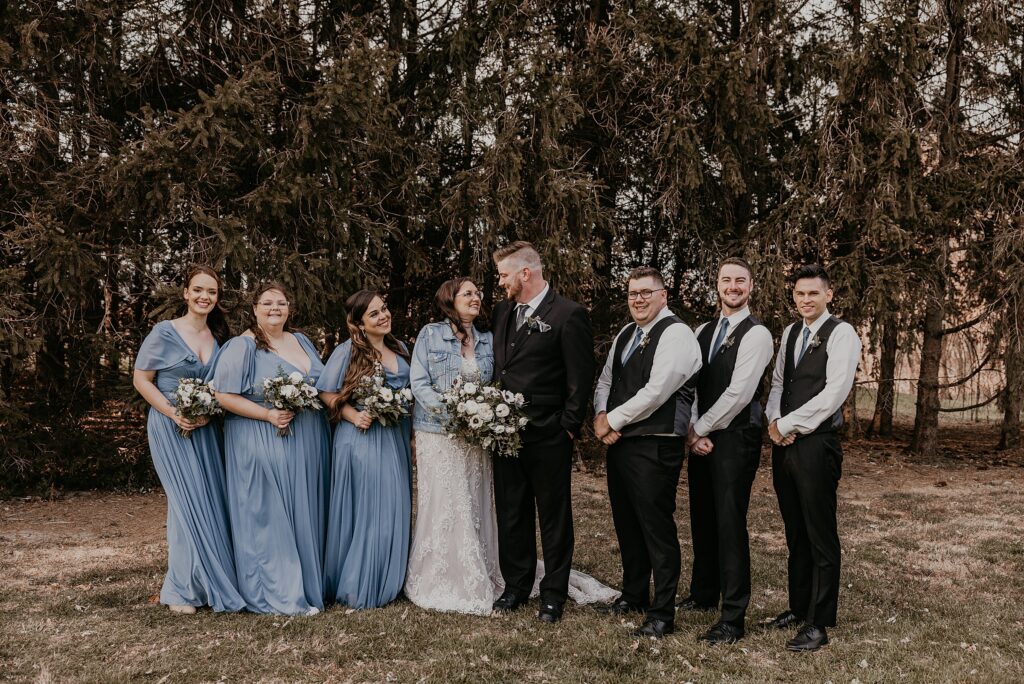 Bridal party portrait at Crescent Hill Acres. Bridesmaids and groomsmen are standing beside the bride and groom. Bridesmaids are in long blue gowns and the bride is wearing a denim jacket. Everyone is laughing. Captured by top Sarnia wedding photographer Ashlee Ellison.
