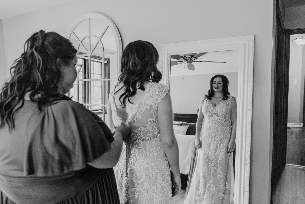 Bride's mother is helping her zip her dress for her Crescent Hill Acres wedding. The image is of the bride's reflection in the mirror. Captured by top Sarnia wedding photographer ashlee ellison.