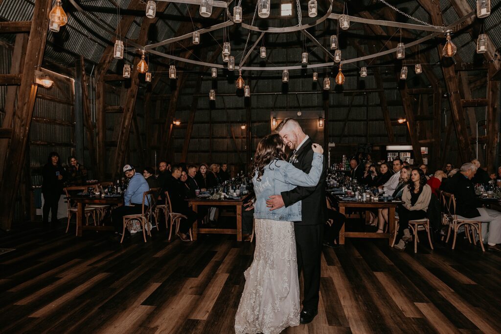 Bride and groom share their first dance in the barn at Crescent Hill Acres. The bride is wearing a denim jacket with her married name on the back. Above them there are vintage edison lights and mason jars with candles. Captured by top sarnia wedding photographer ashlee ellison.