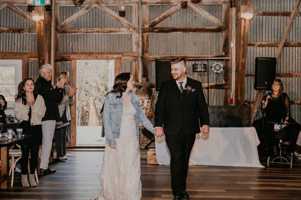 Bride and groom are walking into their reception at Crescent Hill Acres in Plympton-Wyoming. Bride is wearing a denim jacket overtop of her gown. Bride and groom are looking at each other and smiling. Captured by Sarnia wedding photographer Ashlee Ellison.