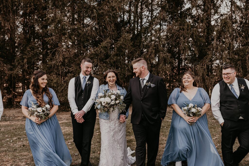 Bride and groom are walking in a line with their wedding party at Crescent Hill Acres. Everyone is laughing. Bride is wearing a denim jacket overtop of her gown. Captured by top Sarnia wedding photographer Ashlee Ellison.